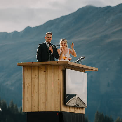 Die Hochzeitsfeier Brautpaar jubelt auf Plattform mit Blumenstrauß in der Hand vor Bergpanorama – Sommerhochzeit und Berghochzeit beim Vermala Wirt im Montafon, Vorarlberg.