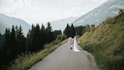 Brautpaar mit traumhaftem Bergblick – Sommerhochzeit beim Vermala Wirt im Montafon Brautpaar spaziert Hand in Hand entlang einer Bergstraße mit Panoramablick auf die Alpen – romantische Sommerhochzeit und Berghochzeit beim Vermala Wirt im Montafon, Vorarlberg.