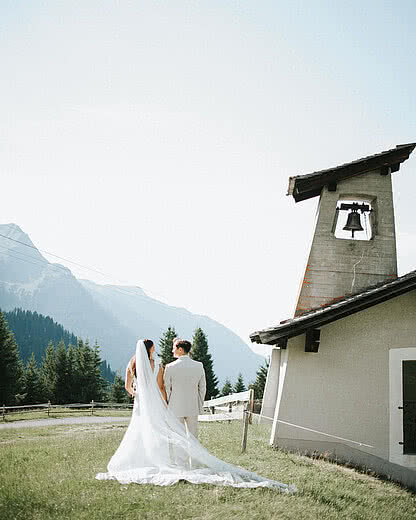 Die Hochzeitslocation Brautpaar geht Hand in Hand zur Kapelle mit Bergkulisse – romantische Berghochzeit und Sommerhochzeit im Montafon beim Vermala Wirt.